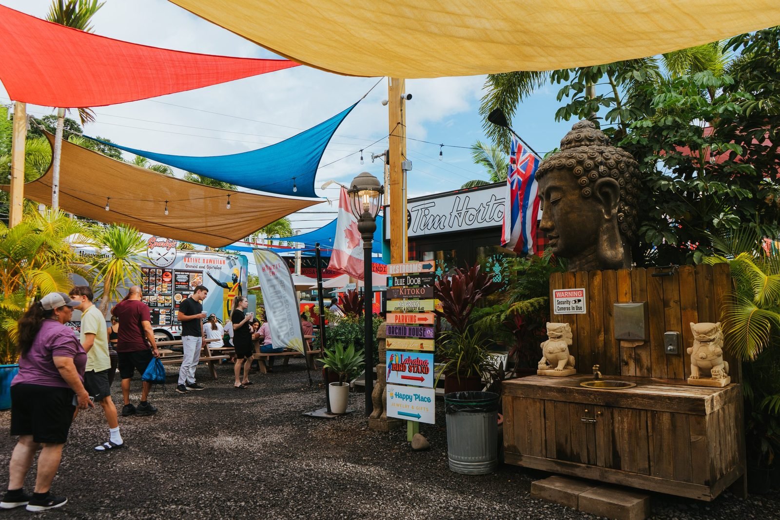 The South Maui Gardens courtyard: colorful shade sails over gravel pathways, a carved stone Buddha, tropical plants, and people relaxing among the food trucks.