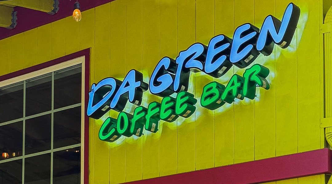 The blue-and-green neon &ldquo;Da Green Coffee Bar&rdquo; sign glowing on the cafe's lime-yellow wall.